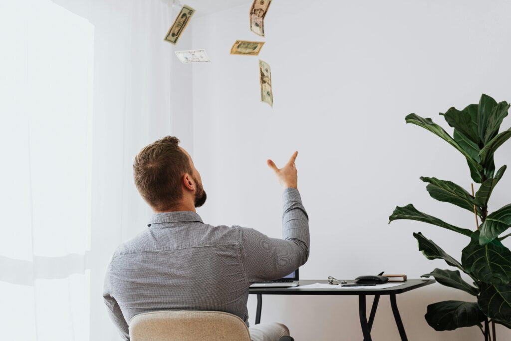 Personal Laons Man throwing cash in the air while sitting at a desk indoors, symbolizing financial success.