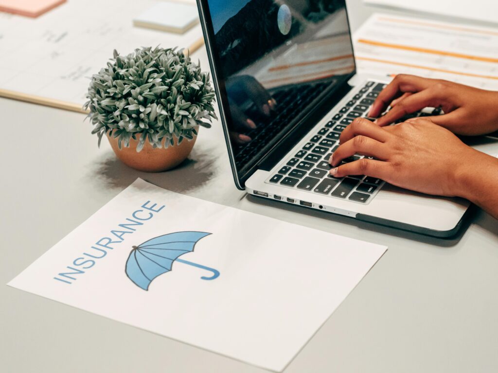 Insurance Hands typing on a laptop at a desk with an insurance paper and plant, suggesting a work environment.
